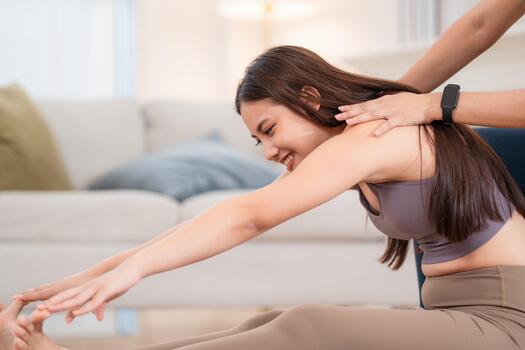 Woman stretches on mat while receiving support from another person in a light room during day photo