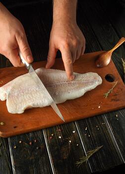 A chef prepares a fresh fish fillet on a wooden cutting board, using a knife with precision. Herbs and spices are scattered around, adding color to the scene photo