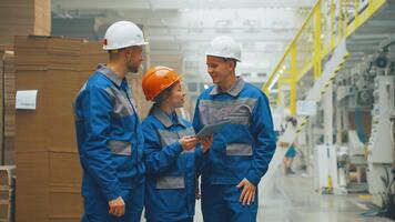 en grupp av ingenjörer och anställda av en kartong förpackning fabrik är talande i de verkstad. fabrik arbetare diskutera de arbete av de transportband och stiga på data in i en digital läsplatta. papper industri. video