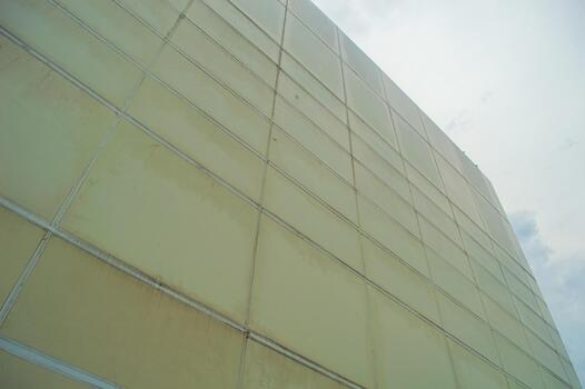 Low angle view of a large, modern building with a pale yellow facade, featuring a grid pattern and visible seams against a cloudy sky, creating an architectural perspective. photo