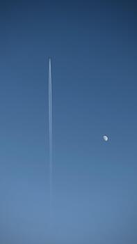 A plane flying in the blue sky and leaving a trace and an incomplete moon visible in the daytime photo