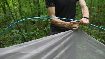 en man försiktigt fäster en tält till en metall ram, säkring varje hörn med precision. omgiven förbi skog grönska, han fokuserar på miljö upp hans skydd för en bekväm stanna kvar i de vild. video