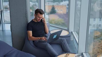 joven masculino persona de libre dedicación utilizando computadora y teléfono a una vez. ocupado Trabajadores por cuenta propia hombre trabajando en un cómodo sitio a el ventana. alto ángulo vista. video