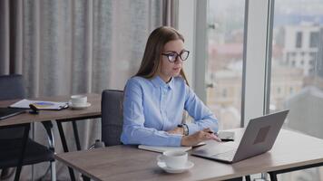 lady arbetssätt i de kontor på de dator. långhårig kvinna tar anteckningar in i papper anteckningsbok och sedan dricka kaffe, ser in i fönster. video
