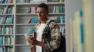 glad afrikansk amerikan man leende studerande bläddring mobil telefon i bibliotek universitet Lycklig etnisk kille manlig använder sig av smartphone uppkopplad studie chattar enhet wiFi internet nätverk bokhyllor bokhandel video