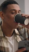 glad nöjd leende afrikansk amerikan man kille manlig bläddring smartphone dricka te kaffe rullning social media Ansökan vilar på soffa Hem använder sig av mobil telefon grej missbrukare användare chattar video