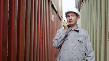 caucasian ingenjör förman i en grå utrusta kommunicerar genom en walkie talkie medan övervaka behållare läser in på en frakt lager, säkerställa slät importera och exportera operationer. video