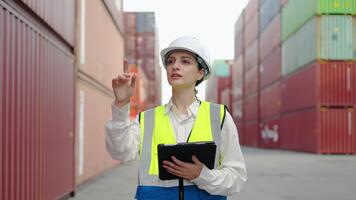 At a busy container yard, a skilled female caucasian engineer in a uniform and white hard hat checks work using a tablet. She ensures safety and efficiency in ongoing operations. video