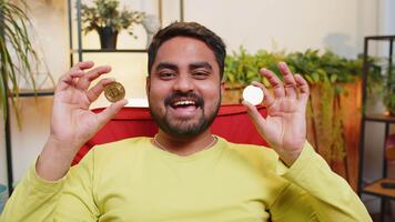 indisch Mann Freiberufler halten zwei Gold Münzen, erfolgreich Entwickler Programmierer. Kerl Lager Händler verdienen Bitcoins nach online Überwachung Handel Operationen. zunehmend Vermögen, finanziell der Wohlstand video