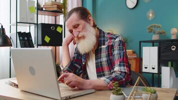 fatigué caucasien Sénior homme ennuyé travail sur portable tandis que séance dans Accueil bureau. somnolent grand-père ne peux pas concentrer sur travail et les types paresseusement. Masculin pigiste est ne pas intéressé dans Faire une projet. video