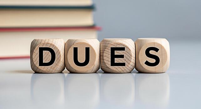 Wooden blocks spelling DUES on a table with books in the background representing financial obligations and responsibilities in an educational or professional setting conveying a sense of commitment photo