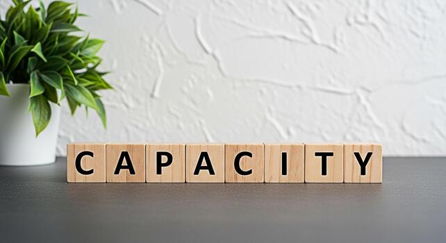 Wooden blocks spelling CAPACITY on a dark surface symbolizing potential and ability in a bright minimalist setting with a green plant representing growth and expansion of skills and resources photo