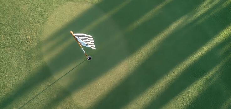 Aerial view of golf flag on green course with long shadows cast by trees. vibrant green grass contrasts beautifully with black and white flag, creating serene golfing atmosphere photo