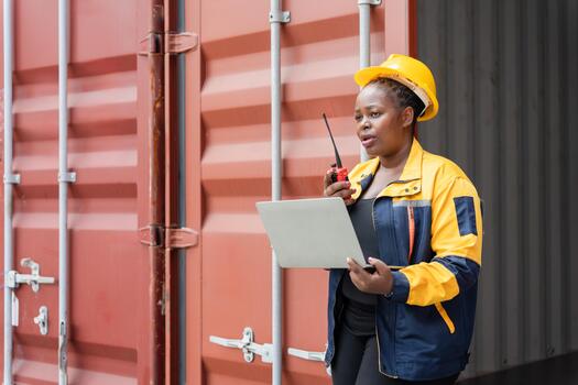 Confident dock worker managing logistics with laptop and radio at shipping terminal, Female industrial worker communicating via walkie talkie while using laptop at container yard photo