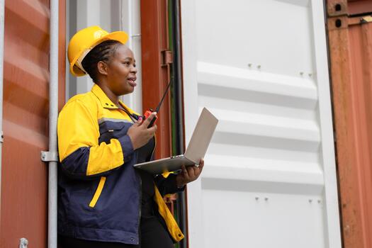 Female industrial worker communicating via walkie talkie while using laptop at container yard, Confident dock worker managing logistics with laptop and radio at shipping terminal photo