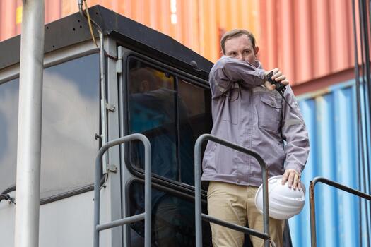 Industrial worker using radio communication at cargo yard, Logistics operator standing on machine platform at shipping port, Safety worker inspecting equipment at container terminal photo