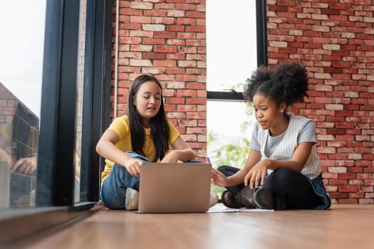 Diverse Students Collaborating on Laptop Homework in a Casual Setting, Teenagers Studying Technology While Sitting on the Floor, Friends Using Computer Near a Window and Brick Wall photo