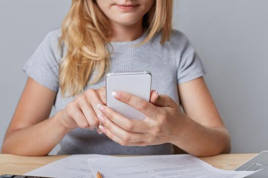 Cropped shot view of blonde female enterpreneur holds smart phone, surrounded with documents, recieves messages, types answers, isolated over grey background. Online communication, networking concept photo