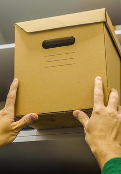 Hands lifting a sealed cardboard storage box onto a shelf, symbolizing home organization, storage solutions, moving tasks, and decluttering routines. High quality photo