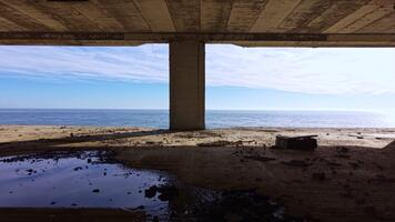 Serene coastal view from beneath an old structured bridge during a calm morning at the beach, offering a glimpse of clear skies and inviting waters video