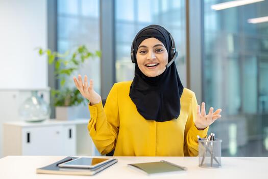 simpático hembra cliente Servicio representante vistiendo auriculares y hijab explicando alguna cosa utilizando gestos mientras trabajando a su escritorio en moderno oficina foto