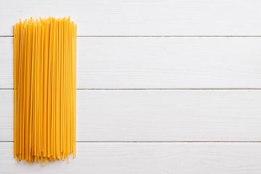 A neat bundle of dry, uncooked spaghetti strands rests vertically on a white, rustic wooden background, leaving ample copy space on the right photo