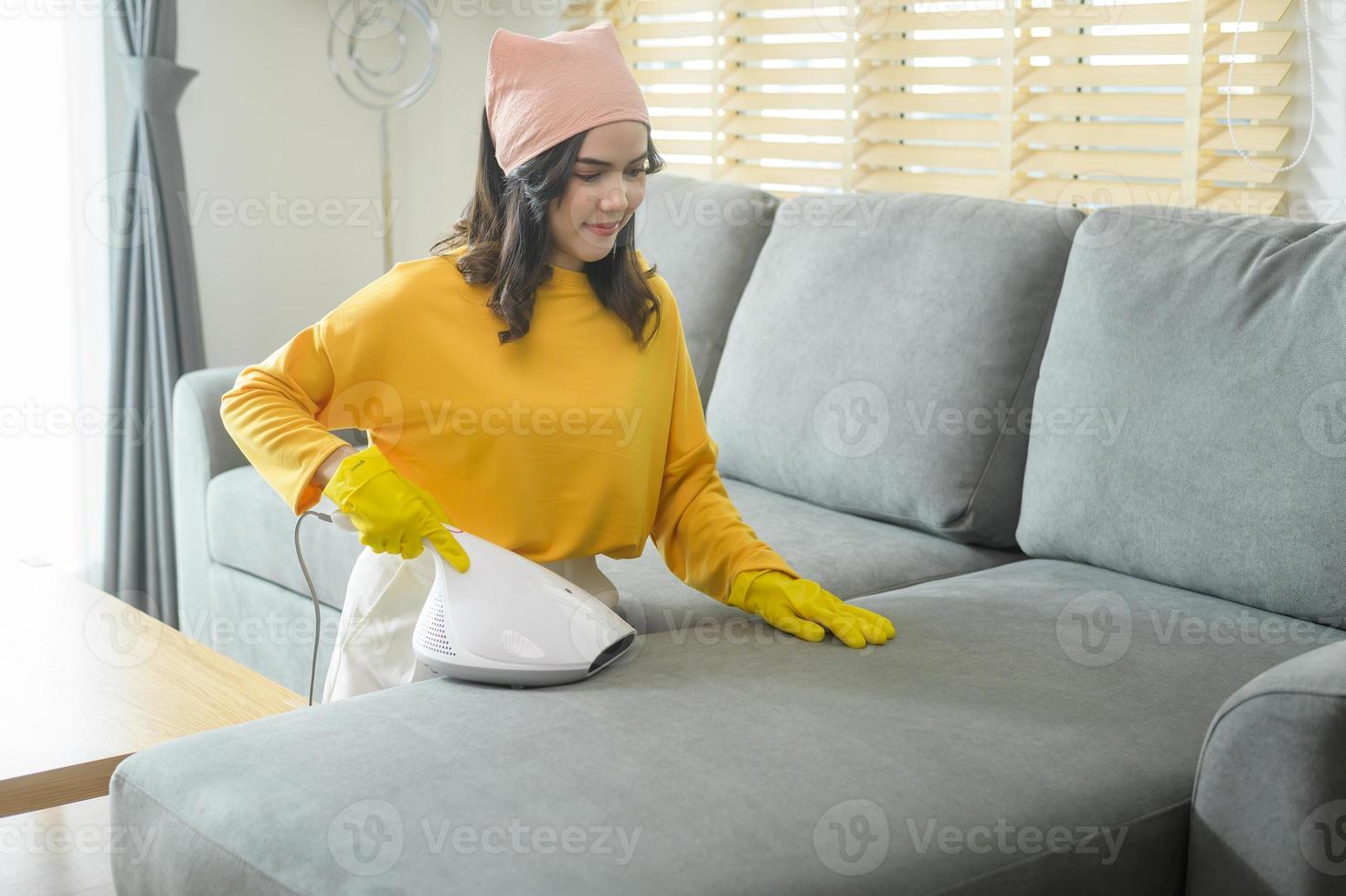 giovane donna felice che indossa guanti gialli e pulisce un divano nel soggiorno con l'aspirapolvere. foto