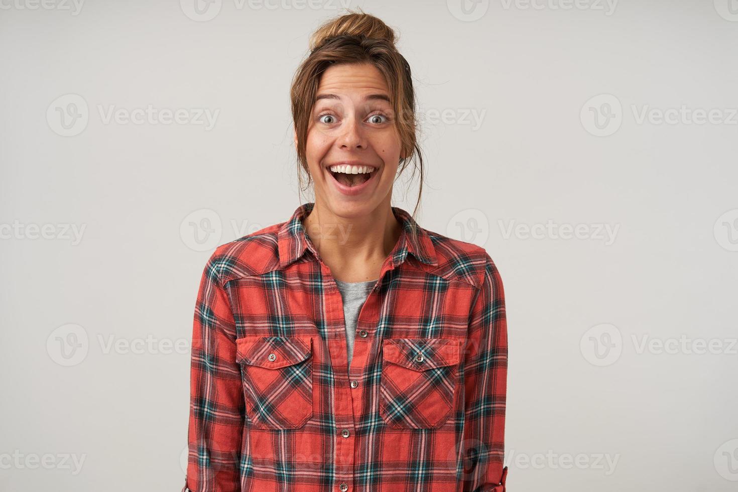 felice giovane donna in camicia a scacchi in piedi su sfondo bianco con le mani in giù, guardando con gioia alla telecamera con la bocca larga aperta, con acconciatura casual e trucco naturale foto