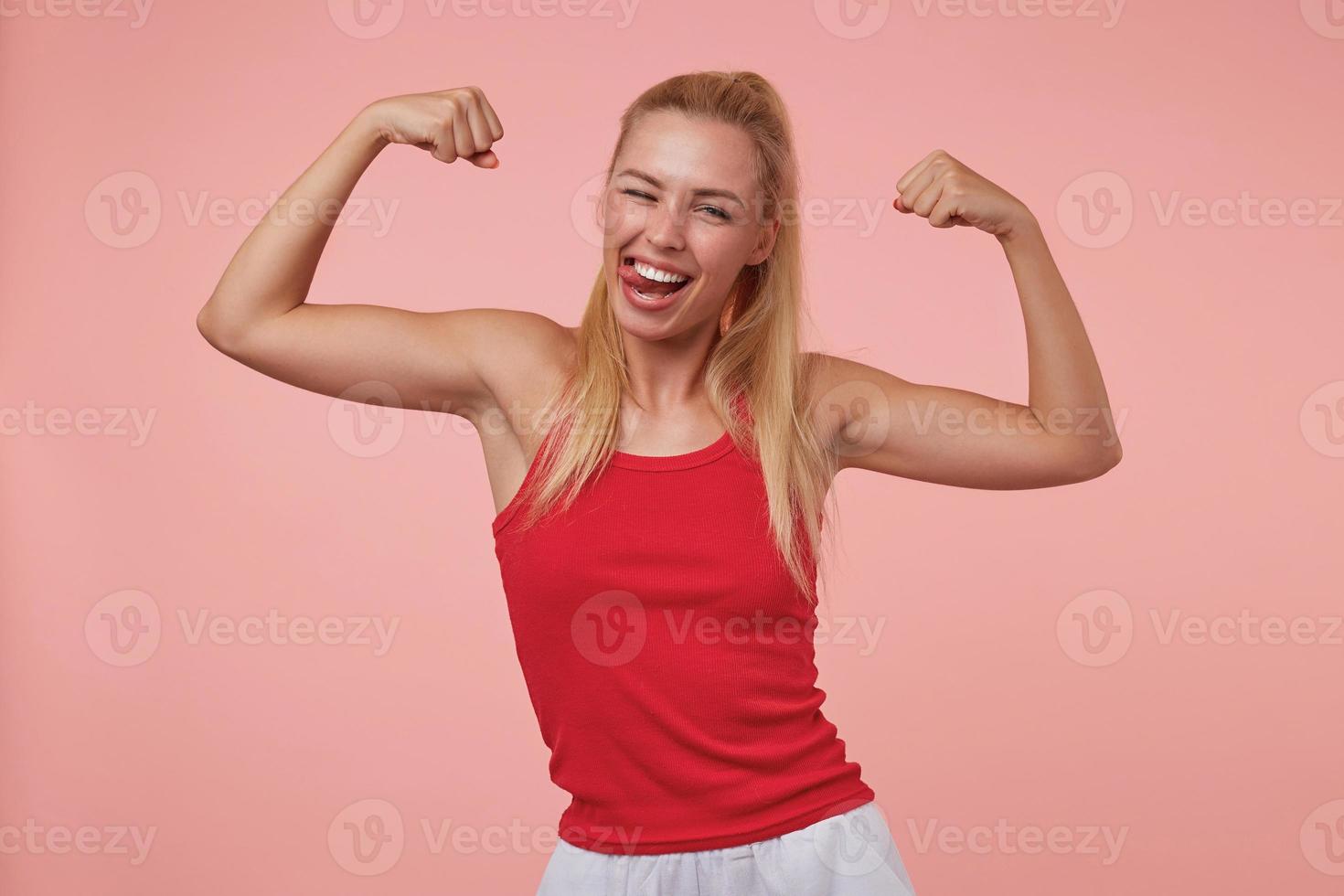 giovane donna allegra e di bell'aspetto con acconciatura casual che mostra le sue mani sportive, sorride ampiamente, mostra la lingua e strizza l'occhio alla telecamera, isolata su sfondo rosa foto