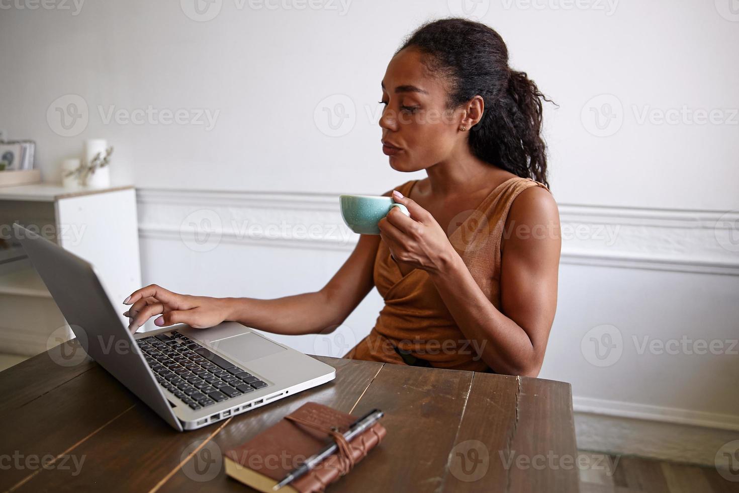ripresa in interni di una bella donna riccia dalla pelle scura che posa sopra lo spazio di lavoro con la faccia seria, bevendo caffè mentre digita le note sul suo laptop foto