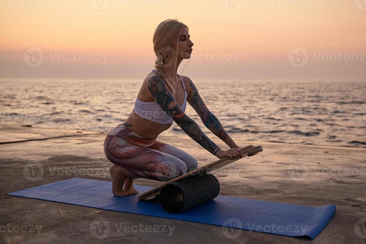 scatto all'aperto di una donna sportiva bionda con un corpo in buone condizioni fisiche in posa con vista sul mare, fare sport al mattino presto con tappetino e tavola per l'equilibrio, indossare top sportivo e leggins foto