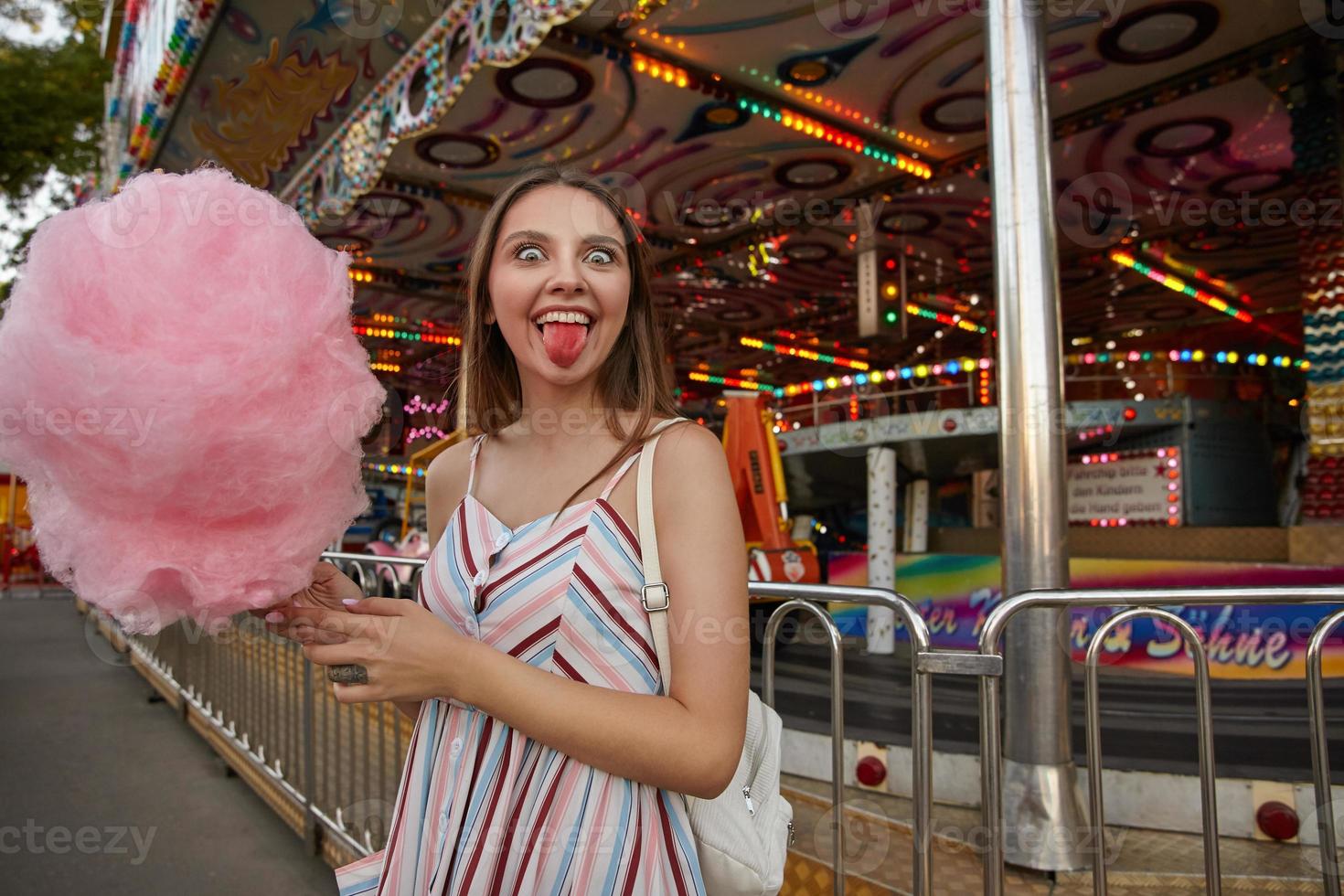 divertente donna bruna con gli occhi aperti con i capelli lunghi in abito estivo leggero in posa sopra il parco divertimenti, guardando la fotocamera felicemente e mostrando la lingua rosa dopo aver mangiato zucchero filato foto