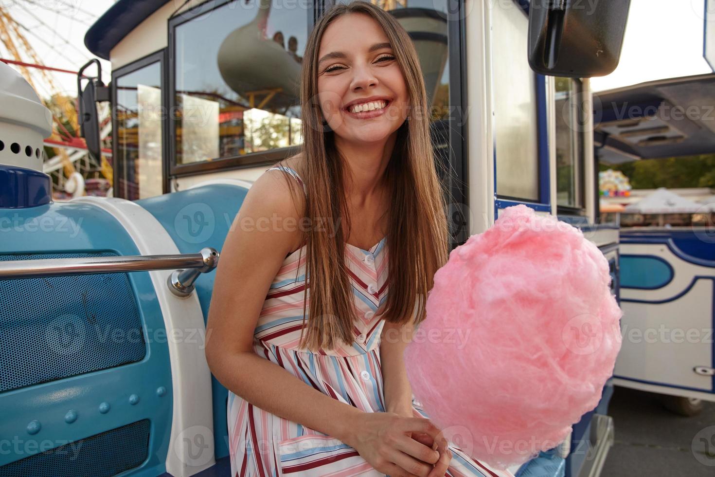 Ritratto di bella giovane donna bruna in abito romantico che guarda la fotocamera con un sorriso affascinante, appoggiandosi a un finto treno a vapore e tenendo in mano zucchero filato rosa foto