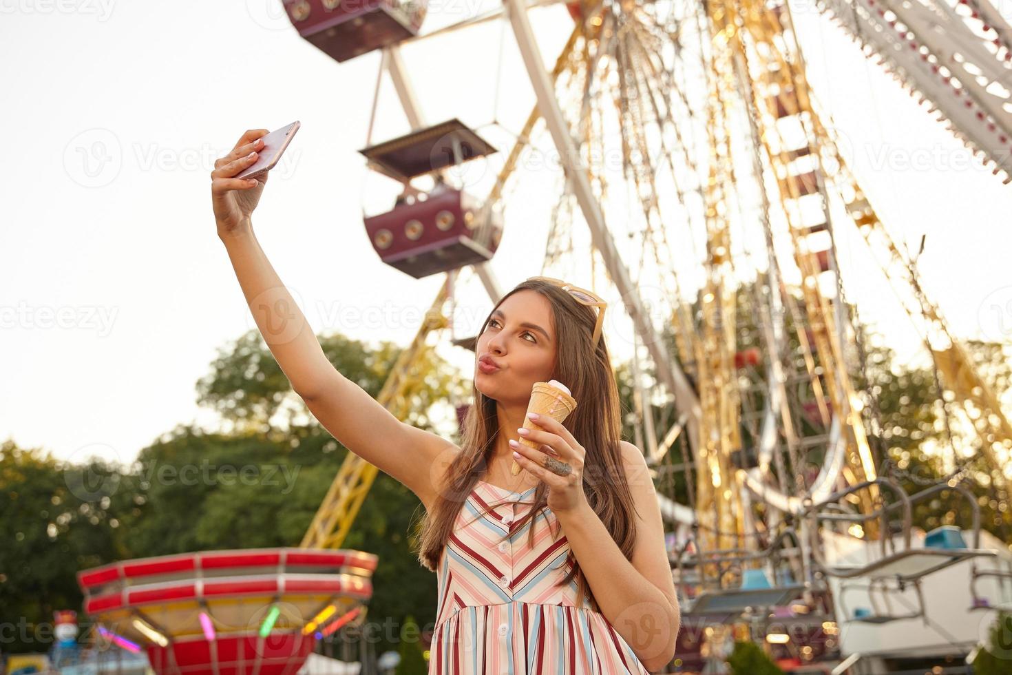 foto all'aperto di una giovane donna dai capelli lunghi in posa sul parco delle attrazioni con le labbra increspate, tenendo in mano un cono gelato e facendo selfie con il suo smartphone