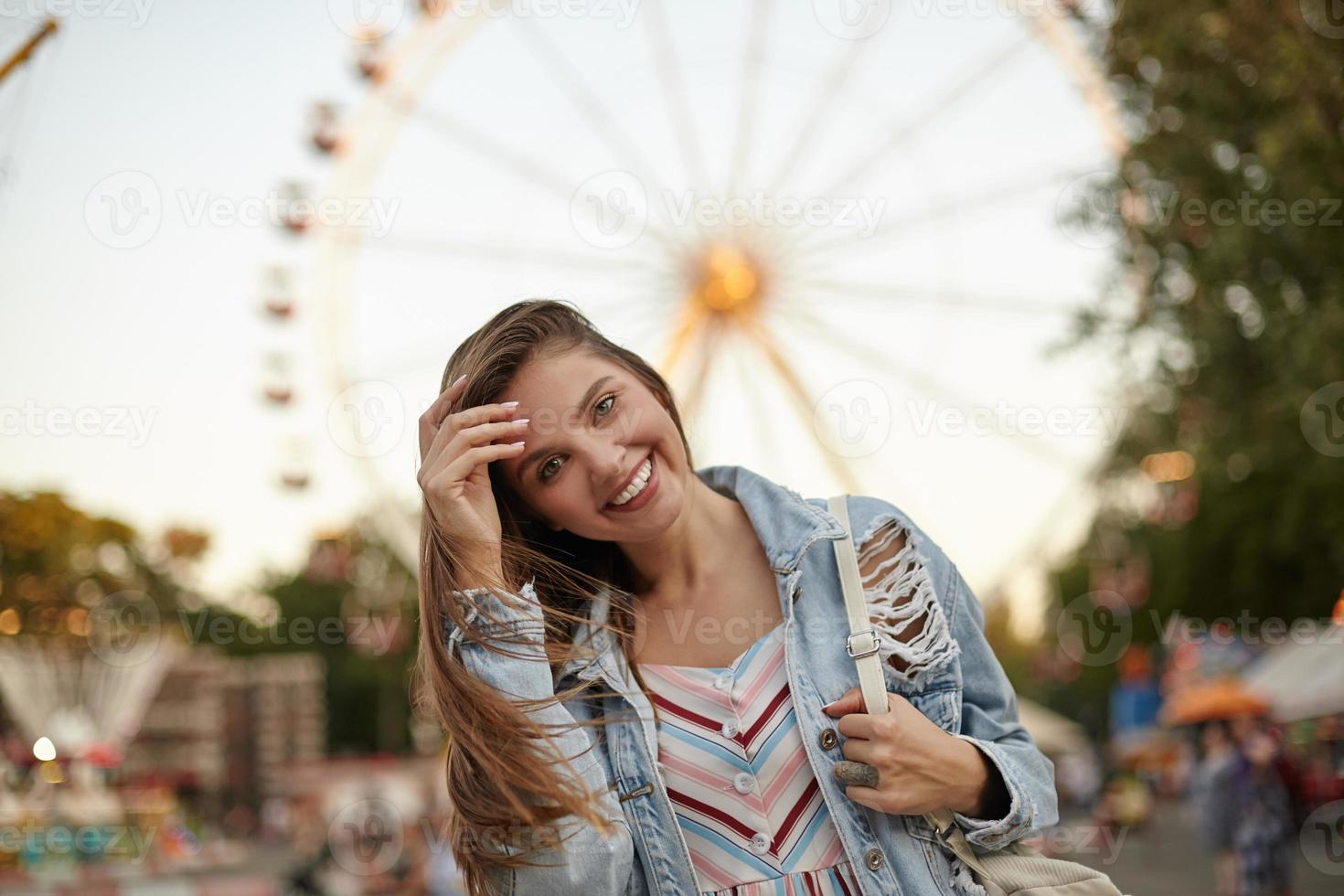 foto all'aperto di bella donna bruna che si raddrizza i capelli e guarda alla fotocamera con un ampio sorriso affascinante, in piedi sopra la ruota panoramica in una calda giornata luminosa in abiti casual