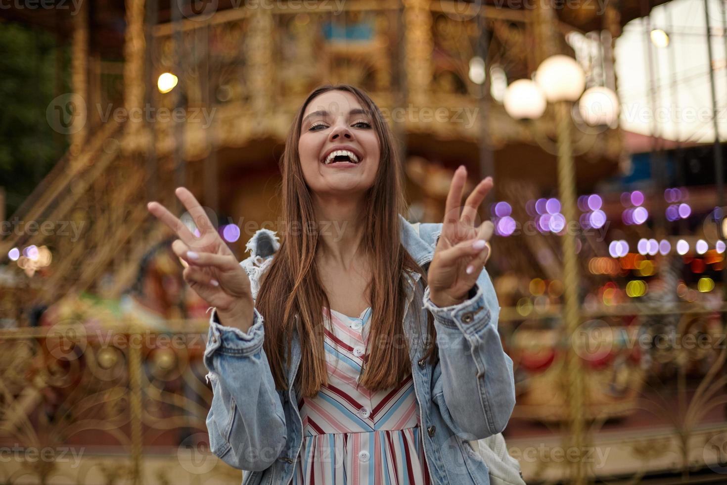 scatto all'aperto di una bella giovane donna con lunghi capelli castani che indossa un abito romantico e un cappotto di jeans, in piedi sopra la giostra nel parco delle attrazioni, sorride alla telecamera e alza le dita nel segno della vittoria foto