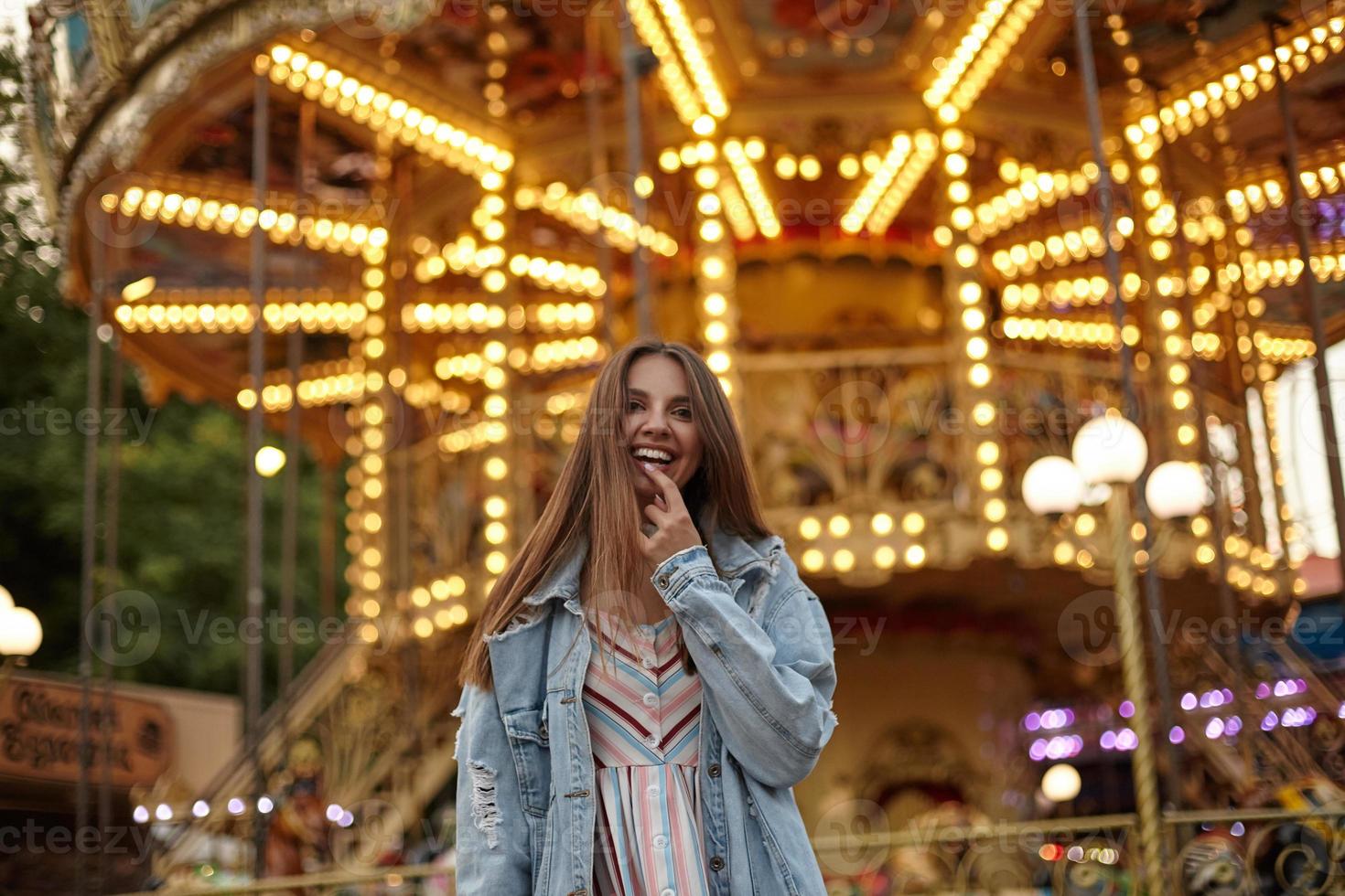 attraente giovane donna bruna dai capelli lunghi che cammina attraverso il parco divertimenti in abito romantico e cappotto di jeans, guardando alla telecamera con un ampio sorriso sincero e tenendo l'indice sul labbro inferiore foto