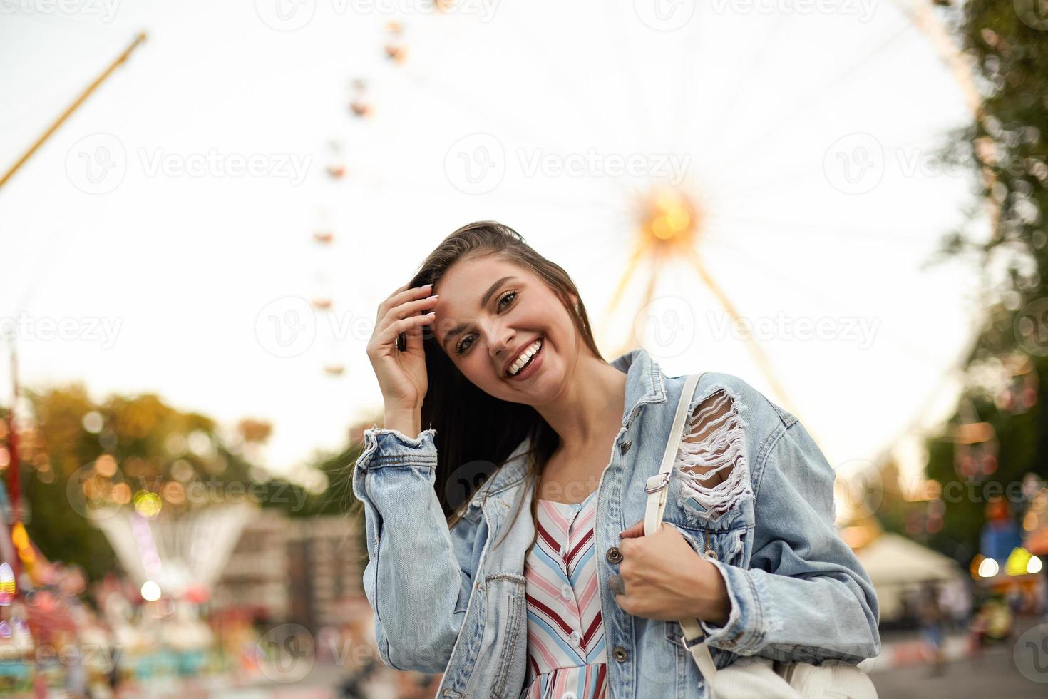 giovane bella bruna allegra femmina in jeans alla moda cappotto in piedi sopra la ruota panoramica nel parco divertimenti, guardando felicemente alla telecamera e raddrizzandosi i capelli foto