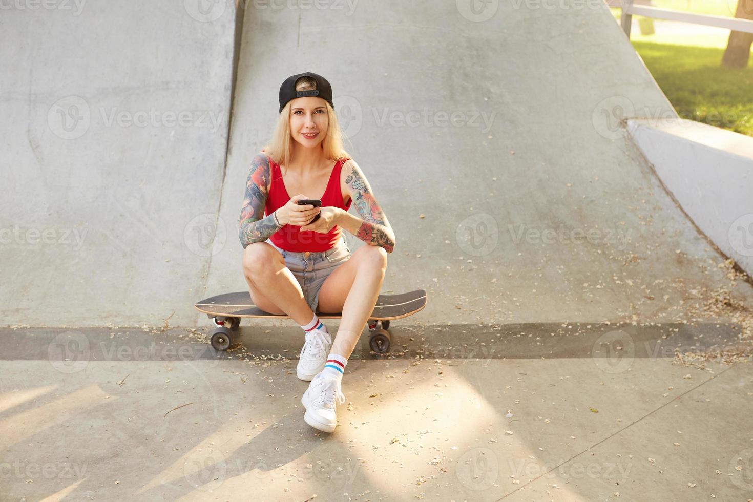 giovane bella donna bionda tatuata in berretto da baseball che guarda alla telecamera in stato d'animo positivo, seduta su skateboard sopra il parco cittadino con lo smartphone in mano foto