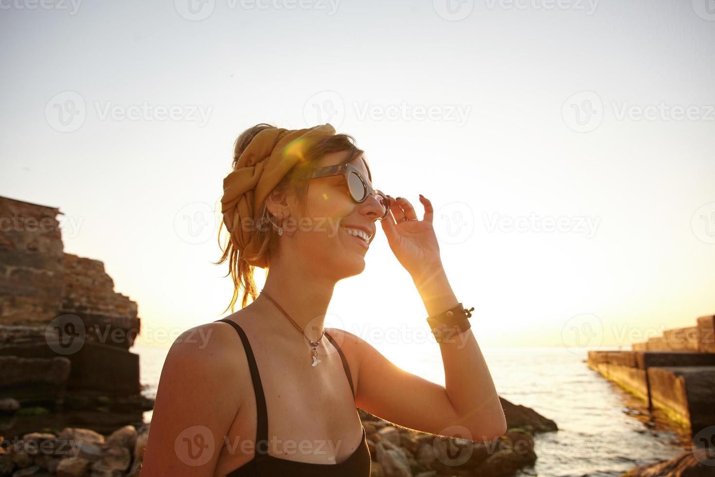 soleggiata foto all'aperto di affascinante giovane donna in fascia in posa sul mare, guardando da parte con un sorriso morbido e toccando i suoi occhiali da sole con la mano