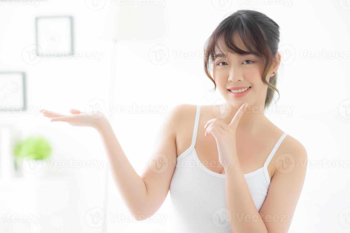 bella giovane donna asiatica che sorride e mostra il prodotto per la cura della pelle per la pubblicità in camera da letto, ragazza asiatica di bellezza che presenta cosmetici per il trucco per la pubblicità con sorpresa sui palmi in camera. foto