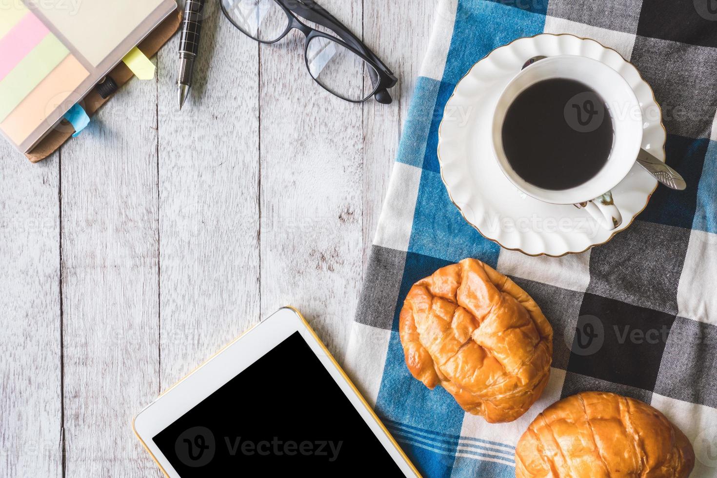 vista dall'alto della tazza di caffè con pane, tovaglia, taccuino, penna, occhiali e tablet su sfondo tavolo in legno foto