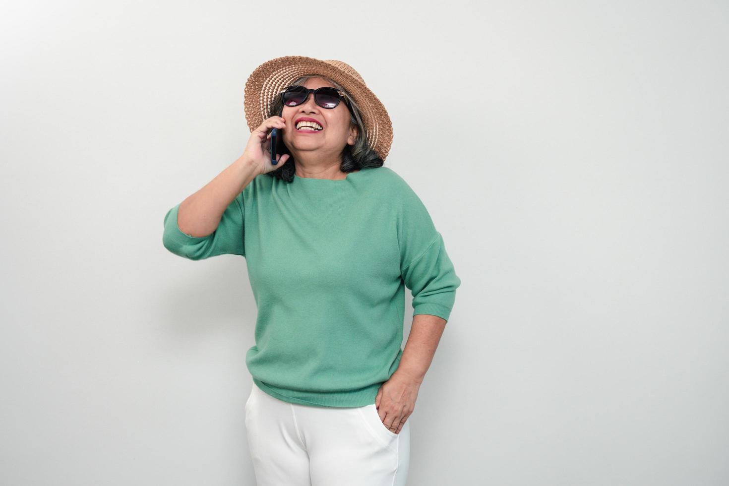 ritratto di donna anziana asiatica felice che indossa occhiali da sole, un cappello e tenendo lo smartphone in piedi isolato su sfondo bianco. concetto di divertimento turistico e divertimento San Valentino dopo il pensionamento foto