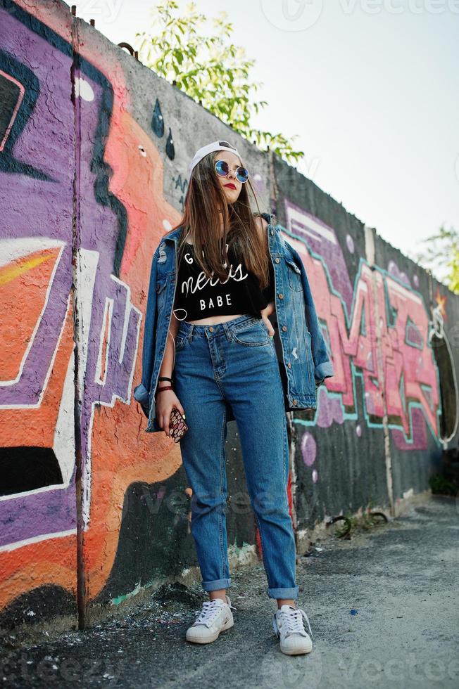 elegante ragazza hipster casual in berretto, occhiali da sole e jeans indossano, ascoltando musica dalle cuffie del telefono cellulare contro il grande muro di graffiti. foto