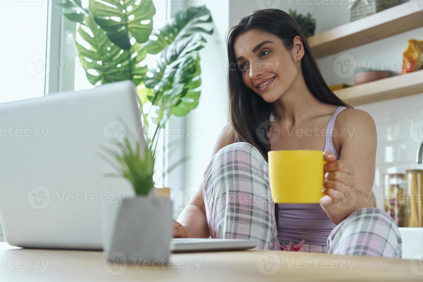 bella giovane donna godendo il caffè del mattino e utilizzando il laptop nella cucina domestica foto