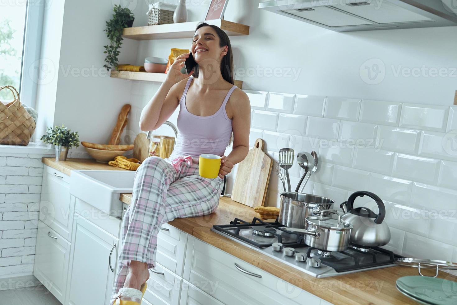 donna felice che parla al cellulare e si gode un caffè mentre si siede sul bancone della cucina foto