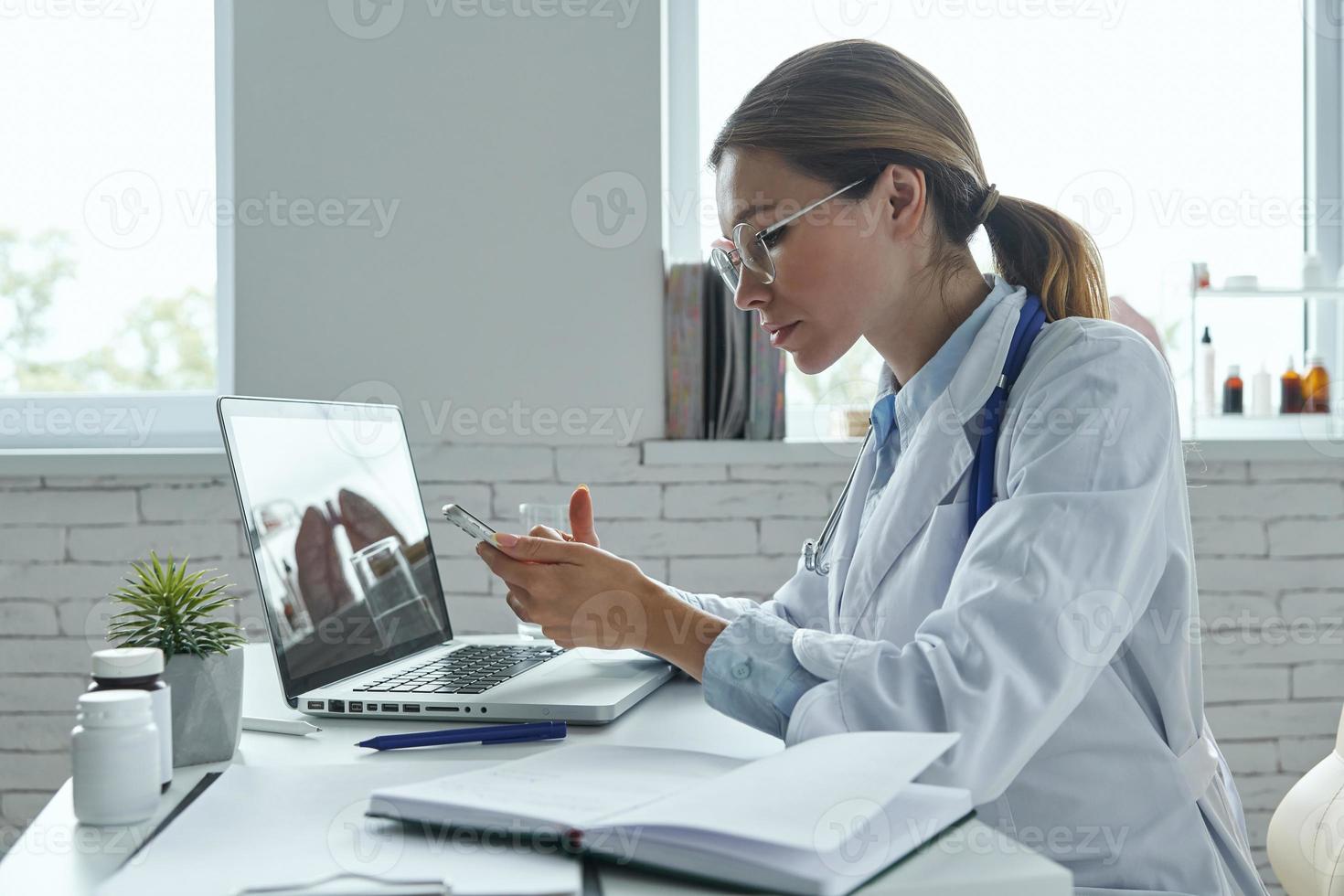 medico femminile sicuro che utilizza lo smartphone mentre è seduto all'ufficio medico foto