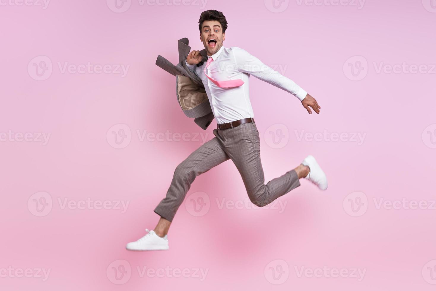 uomo felice in camicia e cravatta che porta giacca sulla spalla mentre salta su sfondo rosa foto