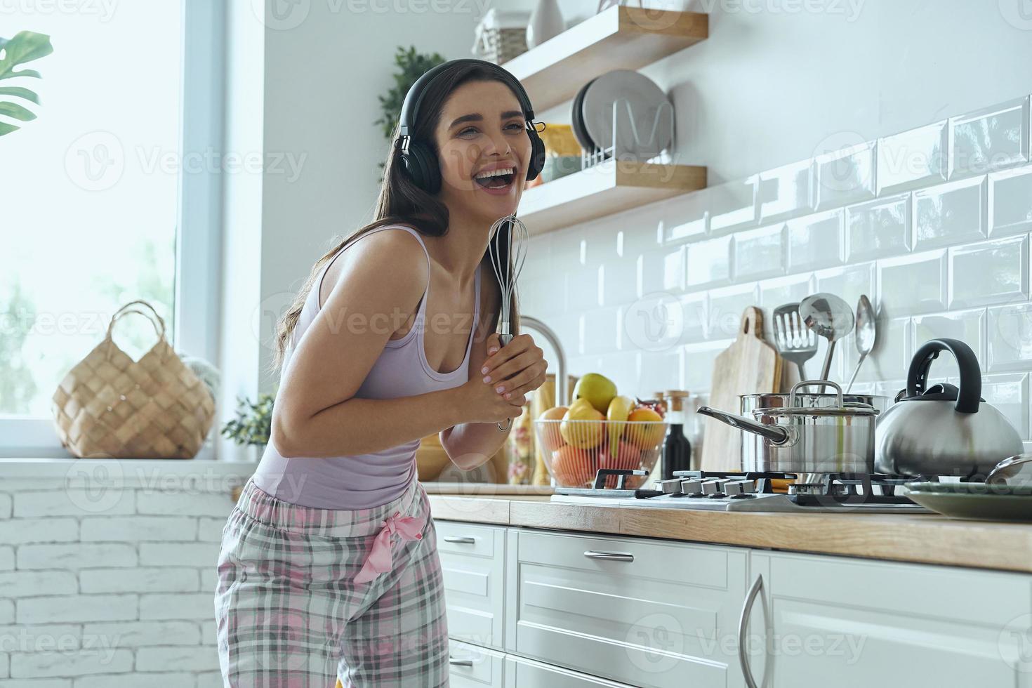 giovane donna giocosa che usa la frusta come microfono e sorride mentre cucina in cucina foto