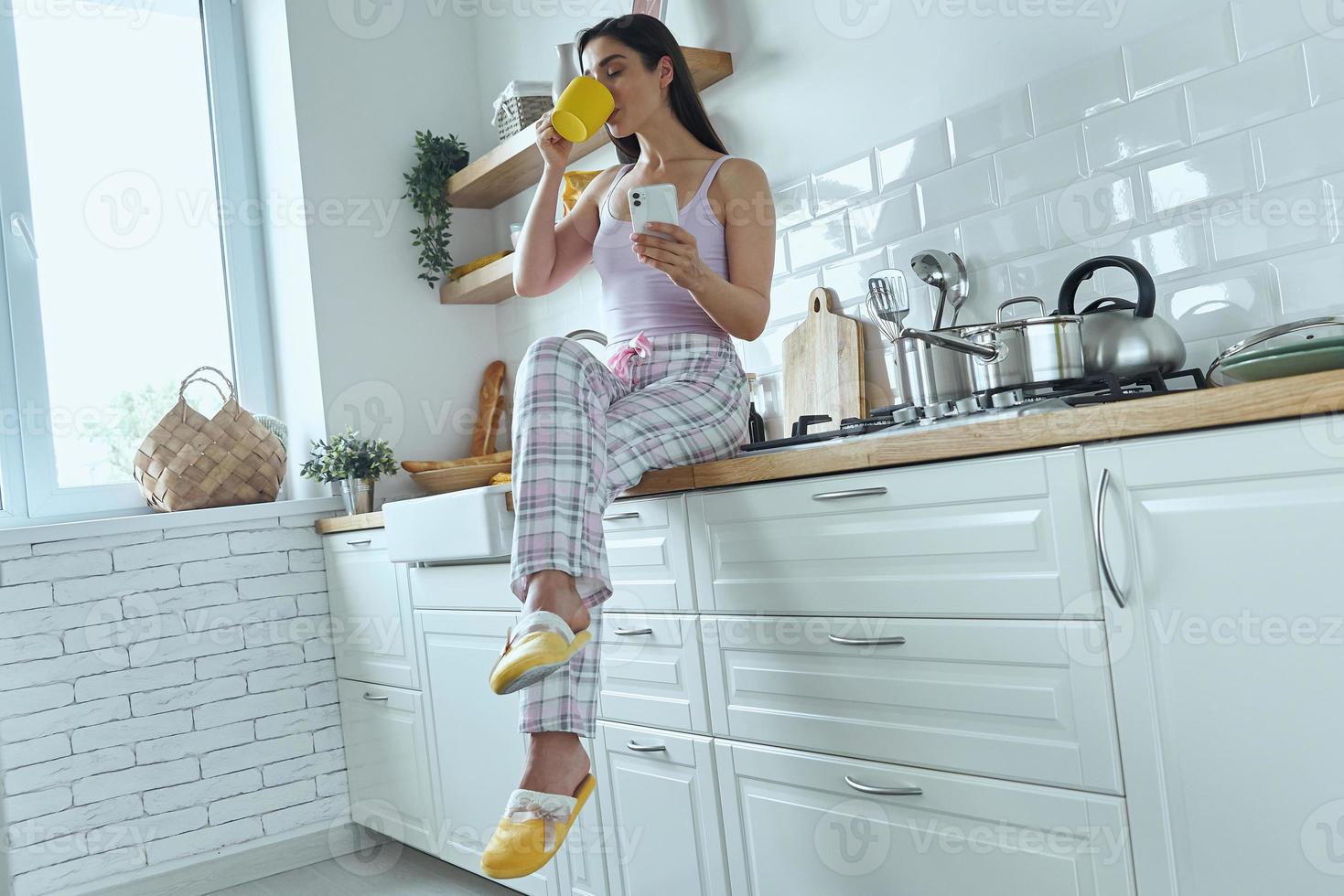 giovane donna in possesso di smart phone e bere caffè seduti sul bancone della cucina foto